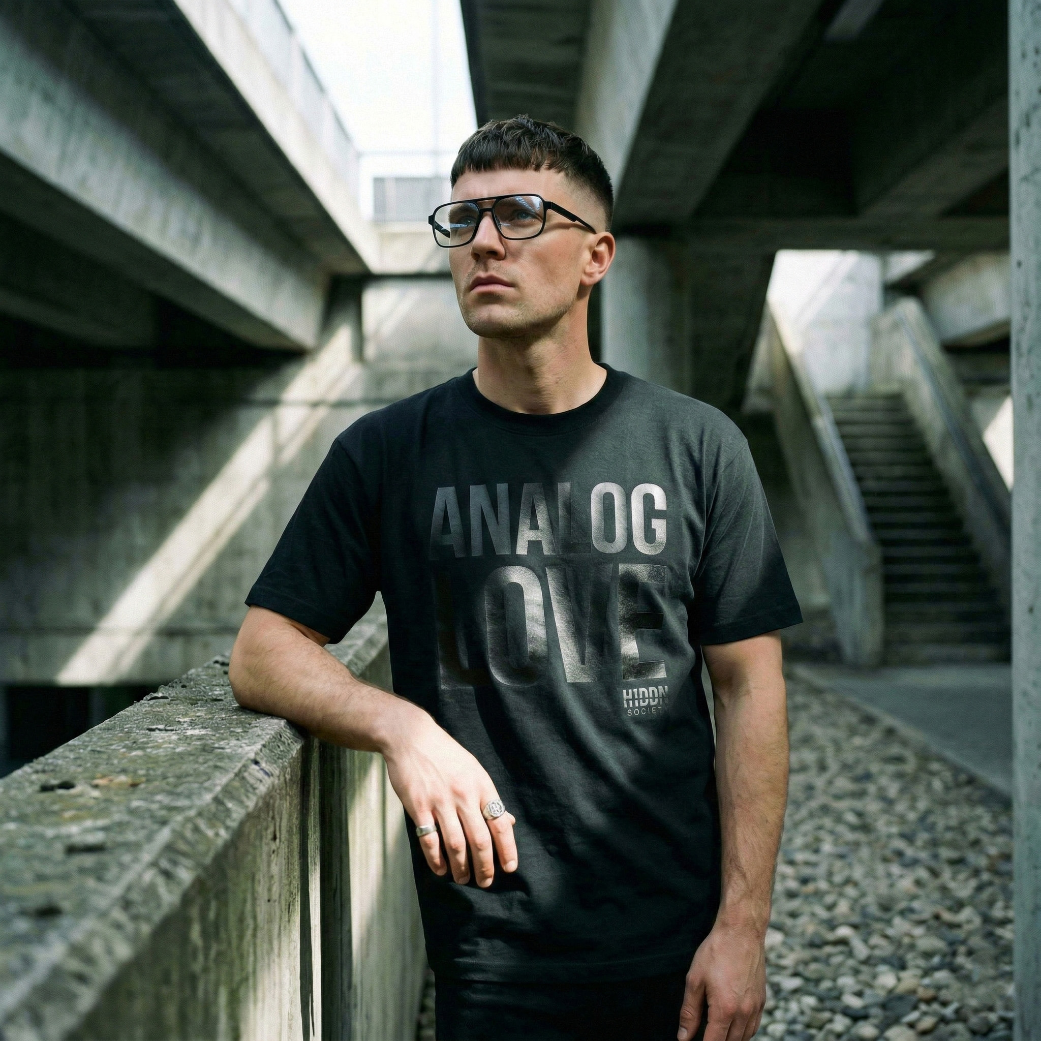 Person wearing a black H1DDN shirt beneath concrete structures